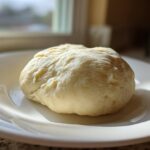A smooth, pale yellow ball of freshly made marzipan recipe dough resting on a white plate.
