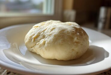 A smooth, pale yellow ball of freshly made marzipan recipe dough resting on a white plate.