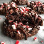 Close-up of fudgy chocolate peppermint cookies, one broken open to show the soft interior, drizzled with chocolate and topped with crushed candy canes.