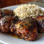 Four pieces of glazed jamaican jerk chicken thighs sprinkled with herbs, served next to a mound of rice on a white plate.