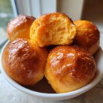 A bowl filled with shiny, glazed sweet potato rolls, one of which is broken open to show the soft, orange interior.