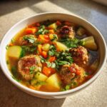 Close-up of a white bowl filled with steaming albondigas soup, featuring meatballs, carrots, potatoes, zucchini, and fresh cilantro.