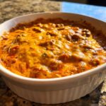 Close-up of a bubbly, baked chili cheese dip topped with melted orange cheddar cheese in a white baking dish.