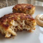 Close-up of a flaky, golden-brown Maryland crab cake with a bite taken out, served next to a dollop of sauce.