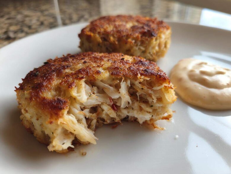 Close-up of a flaky, golden-brown Maryland crab cake with a bite taken out, served next to a dollop of sauce.