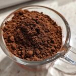 Close-up of rich brown powder, representing homemade hot cocoa mix, filling a clear glass measuring cup.