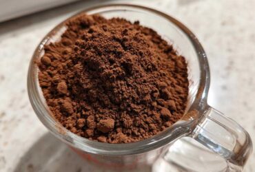 Close-up of rich brown powder, representing homemade hot cocoa mix, filling a clear glass measuring cup.