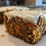 Close-up of a moist slice of carrot cake bars showing shredded carrots and nuts, topped with thick cream cheese frosting.