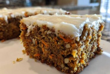 Close-up of a moist slice of carrot cake bars showing shredded carrots and nuts, topped with thick cream cheese frosting.