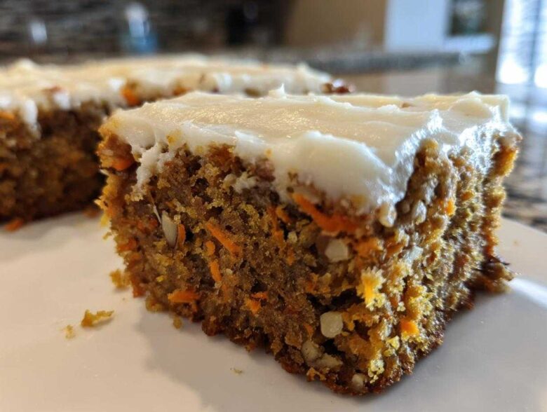 Close-up of a moist slice of carrot cake bars showing shredded carrots and nuts, topped with thick cream cheese frosting.