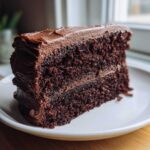 A close-up of a decadent slice of moist chocolate cake recipe with thick chocolate frosting layers.