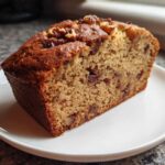 Close-up of a moist slice of date nut bread topped with walnuts, served on a white plate.