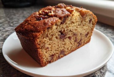 Close-up of a moist slice of date nut bread topped with walnuts, served on a white plate.
