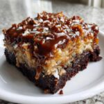 Close-up of a moist slice of german chocolate poke cake featuring a rich chocolate base and thick coconut-pecan topping drizzled with caramel.