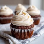 A close-up of one perfectly frosted gingerbread cupcake with creamy white frosting, resting on a striped cloth.