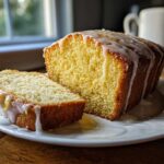 A moist lemon loaf, partially sliced, featuring a thick, white glaze dripping down the sides, perfect for a sweet treat.