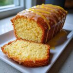 A moist lemon loaf, partially sliced, drizzled with a thick glaze and topped with lemon zest strips.