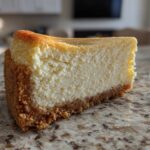 A thick, creamy slice of New York cheesecake with a golden-brown graham cracker crust on a speckled countertop.