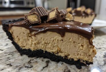 Close-up of a thick slice of no-bake peanut butter pie with a dark crust, peanut butter filling, chocolate topping, and peanut butter cups.