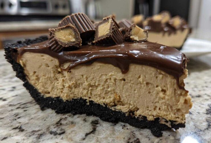 Close-up of a thick slice of no-bake peanut butter pie with a dark crust, peanut butter filling, chocolate topping, and peanut butter cups.
