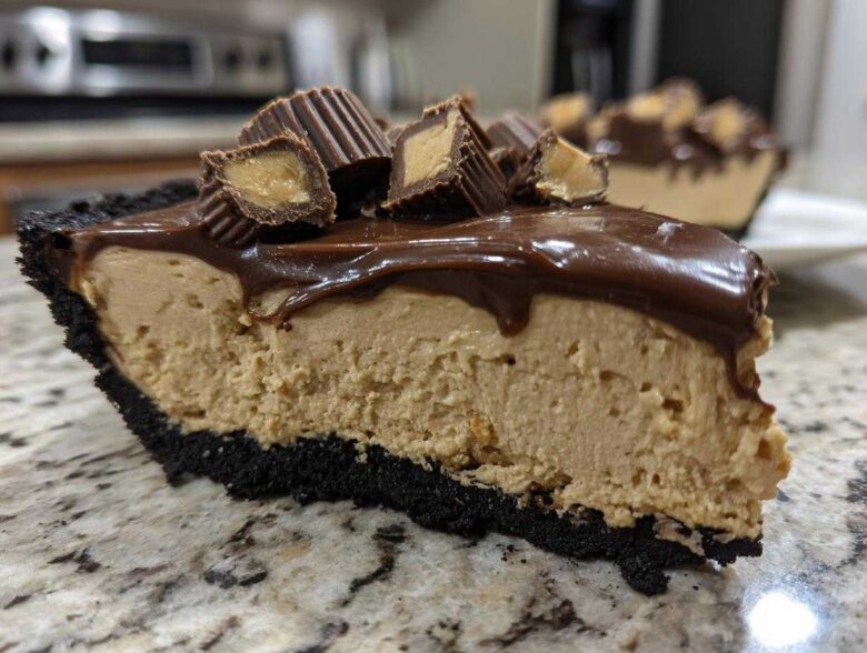 Close-up of a thick slice of no-bake peanut butter pie with a dark crust, peanut butter filling, chocolate topping, and peanut butter cups.