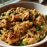 A close-up of creamy, savory one pot chicken and rice mixed with bright green peas, served in a white bowl.