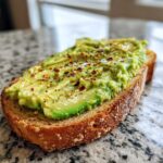 Close-up of a slice of avocado toast, topped with mashed avocado and red pepper flakes.