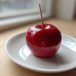 A single, glossy red candy apple with a wooden stick, resting on a white plate on a light wooden surface.