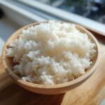 Close-up of perfectly cooked, glossy sushi rice served in a small wooden bowl near a window.
