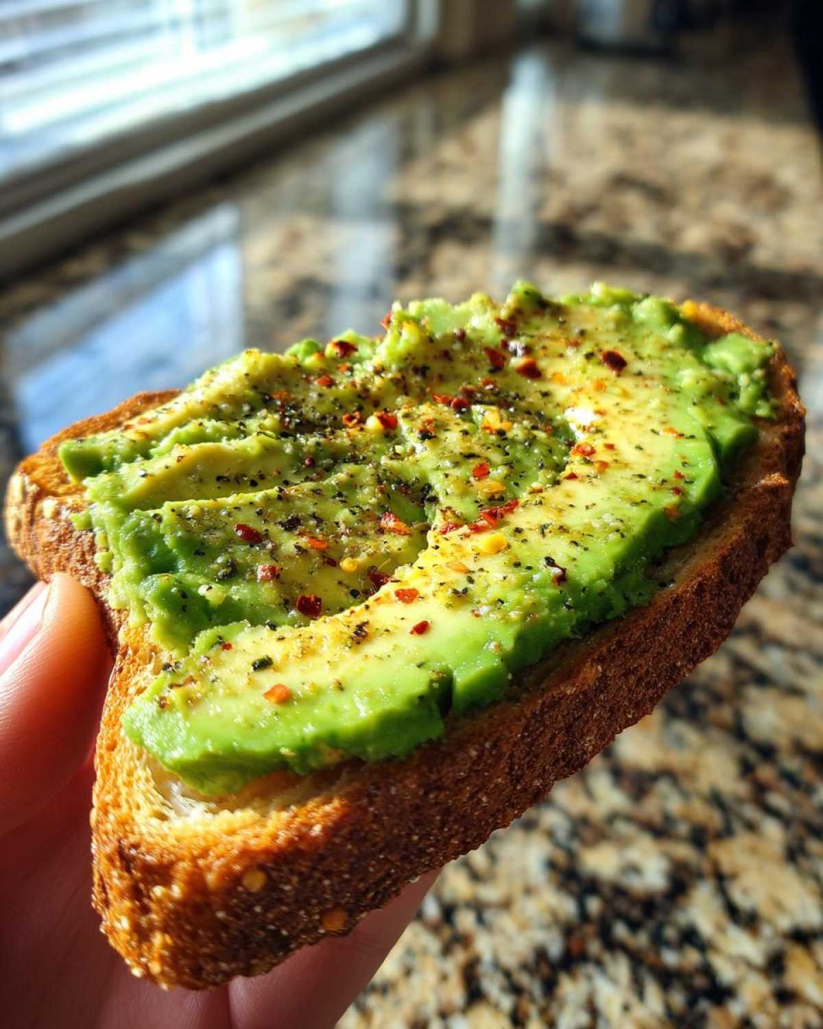 A hand holding a slice of perfectly toasted bread topped with mashed avocado, black pepper, and red pepper flakes for delicious avocado toast.