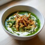Close-up of a bowl of vibrant green Persian noodle soup topped with white beans, yogurt swirl, and crispy fried onions.