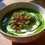 A close-up of a bowl of bright green Persian noodle soup topped with yogurt, herbs, and crispy fried onions.
