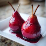 Two glistening, deep red poached pears with stems intact, sitting in a pool of dark syrup on a white dessert plate.