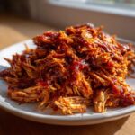 Close-up of a generous serving of saucy, tender pulled chicken piled high on a white plate.