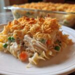 A serving of creamy chicken noodle casserole topped with crispy fried onions sits on a white plate, with the baking dish visible in the background.