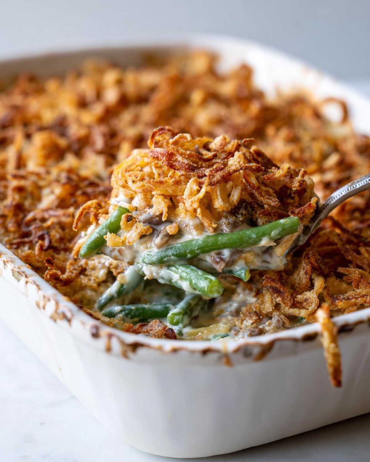 A spoonful of creamy green bean casserole topped with crispy fried onions being lifted from a white baking dish.