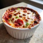 Close-up of a bubbling, baked pizza dip topped with melted cheese, pepperoni, and green peppers in a white ramekin.