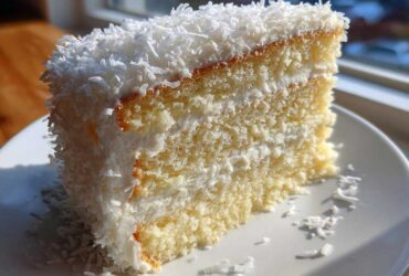 Close-up of a moist slice of coconut cake recipe, layered with white frosting and covered in shredded coconut.