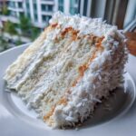 Close-up of a moist slice of white coconut cake recipe layered with frosting and covered in shredded coconut.