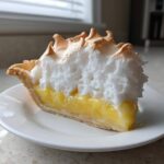 A tall slice of lemon meringue pie showing bright yellow filling and fluffy, toasted meringue.