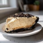 A generous slice of no bake peanut butter pie featuring a thick, creamy filling and chocolate cookie crust, topped with cookie crumbles.