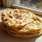 A stack of freshly made, soft corn tortillas with light brown toasted spots resting on a countertop near a window.