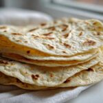 A stack of freshly made, soft corn tortillas with light brown char marks resting on a white cloth.