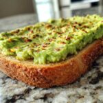 Close-up of creamy avocado toast topped with red pepper flakes, sitting on a granite countertop.