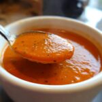 Close-up of a spoonful of thick, orange-red roasted tomato soup being lifted from a white mug.