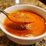 A spoonful of vibrant, creamy roasted tomato soup, garnished with black pepper or herbs, lifted from a white bowl.