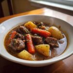 A close-up of a steaming white bowl filled with rich, dark crockpot beef stew featuring chunks of beef, carrots, and potatoes.