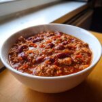 A close-up of a steaming white bowl filled with thick, rich high protein chili featuring ground meat and kidney beans.