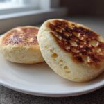 Two golden-brown homemade English muffins showing characteristic holes, resting on a white plate.