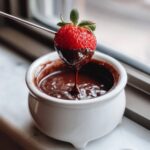 A fresh strawberry being dipped into rich, melted chocolate fondue using a metal skewer.